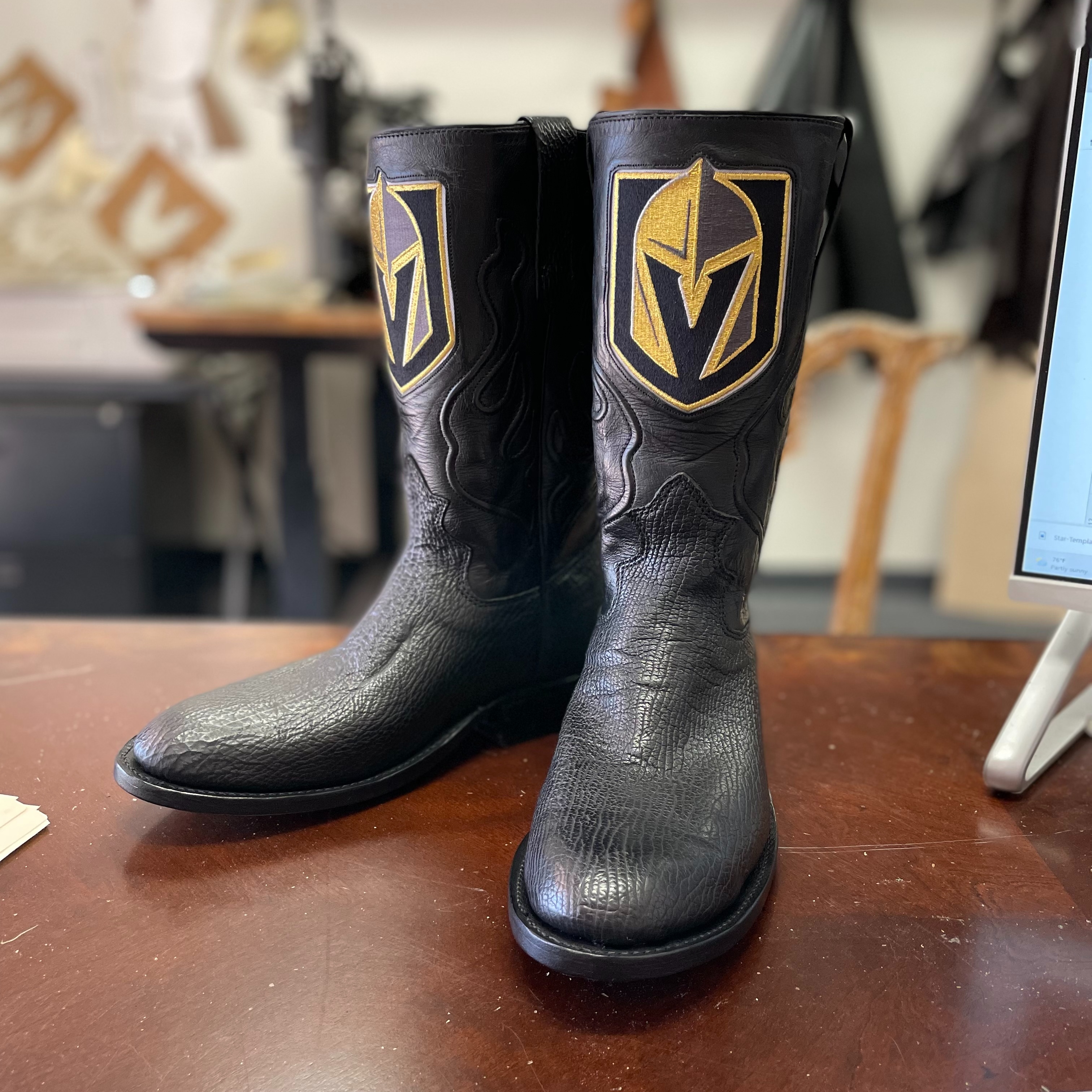 A pair of black custom cowboy boots featuring gold shield embroidery and detailed leather stitching. 