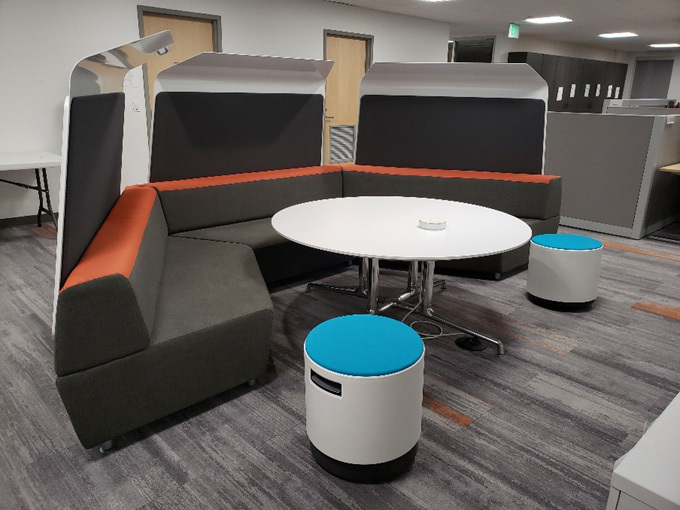 A modern and cozy office lounge area featuring a U-shaped sectional sofa with dark gray cushions and orange accents. The seating arrangement surrounds a round white table on a metallic base, creating a collaborative space for informal meetings or discussions. The setup includes two small, portable stools with white bases and bright blue cushions for additional seating. High-backed partitions provide privacy, and the area is set on contemporary gray and orange-patterned flooring.