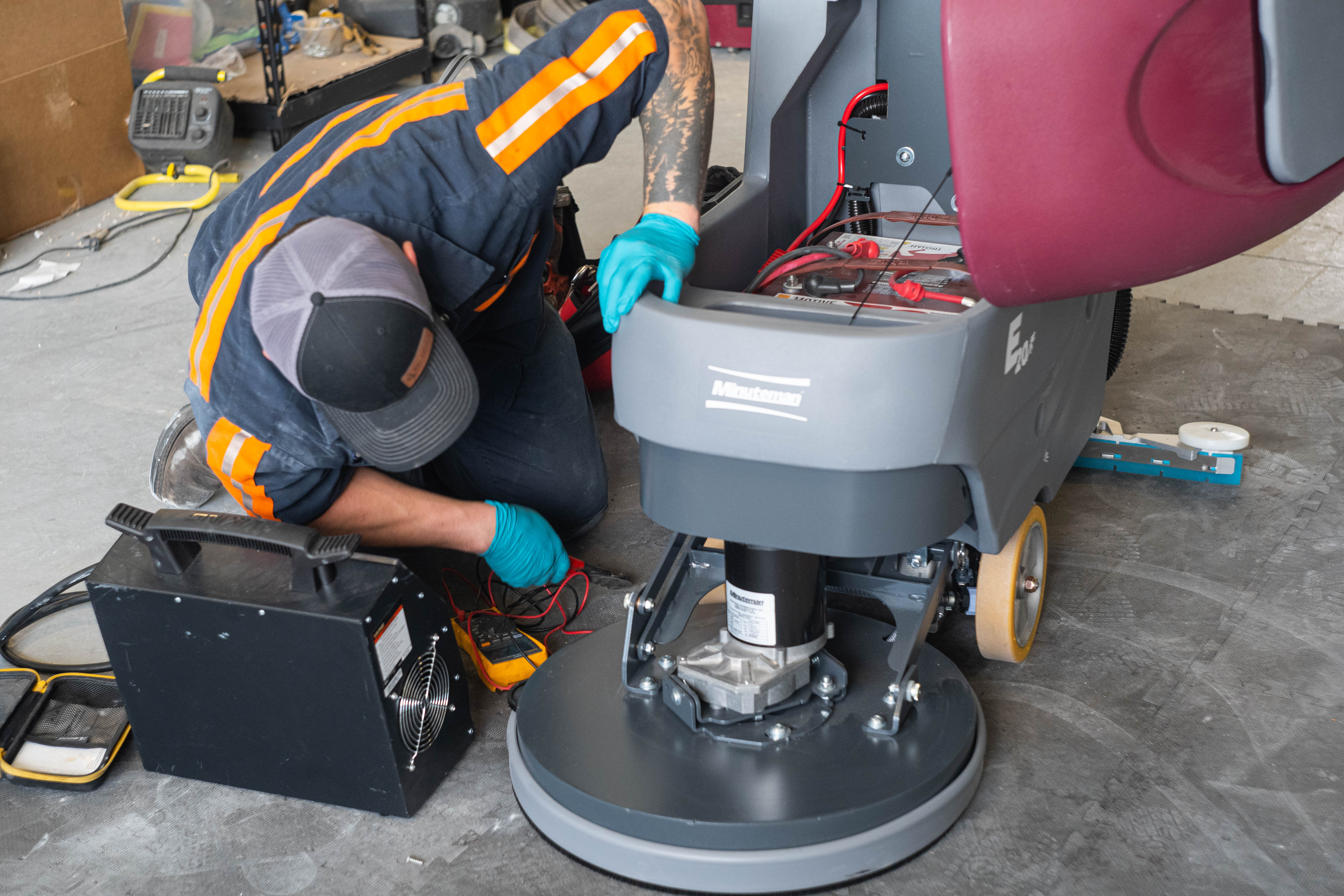 A WaySolutions field technician kneels to perform deep electrical diagnostics on the lower scrub motor of a Minuteman E20 walk-behind floor scrubber. Utilizing precision tools like a digital multimeter alongside a Viking load tester, our Layton-based team expertly traces faults and executes deep mechanical overhauls for heavy-duty commercial fleets.