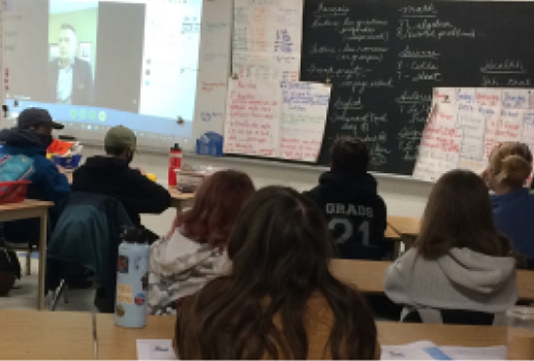 Students sit facing a large classroom screen displaying a virtual speaker. The background shows a blackboard with colorful notes, suggesting a learning environment.