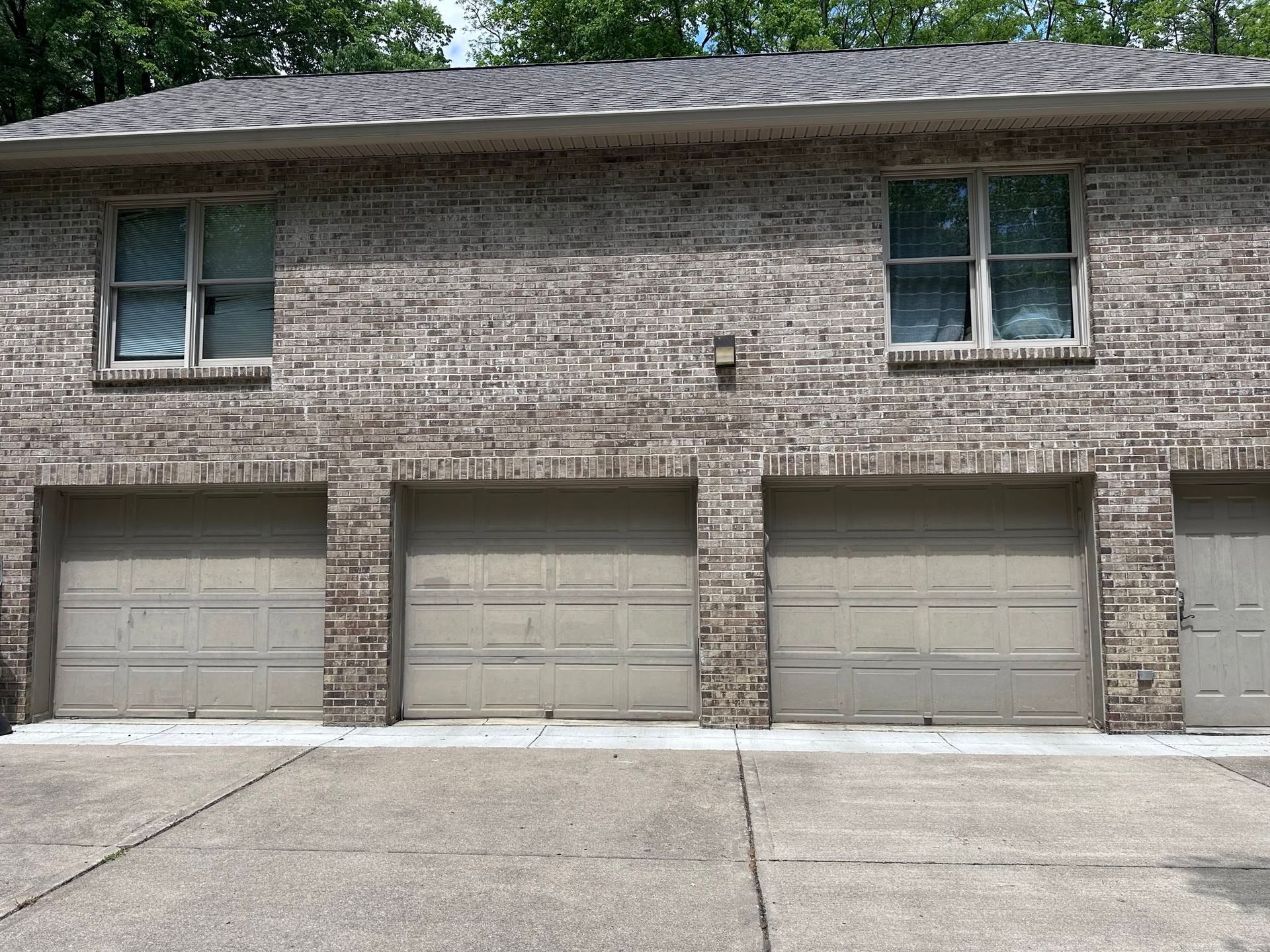 A brick residential building features multiple older gray sectional garage doors with recessed panels, narrow upper sections, concrete aprons, and mature trees surrounding the property for established character and charm.