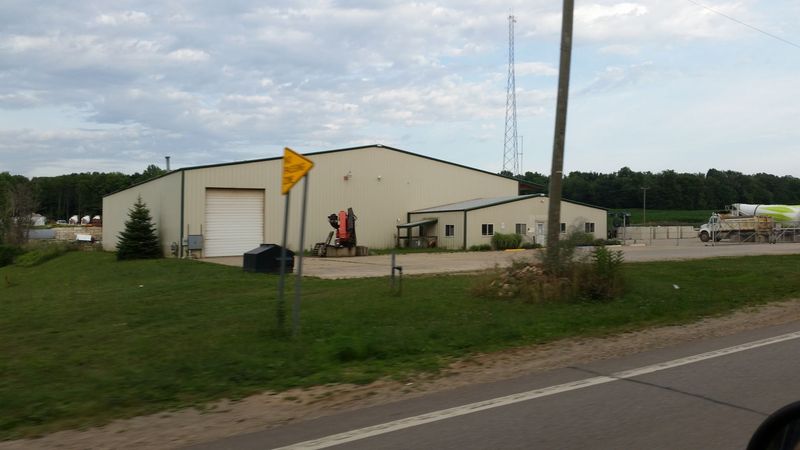 A beige industrial warehouse with a large roll-up door stands on a grassy lot next to a road. A communication tower and a partial view of a cement truck are visible.