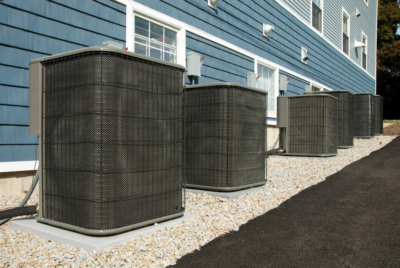 A line of four large, dark grey air conditioning units sits on a concrete pad surrounded by light grey gravel next to a two-story building with blue siding.