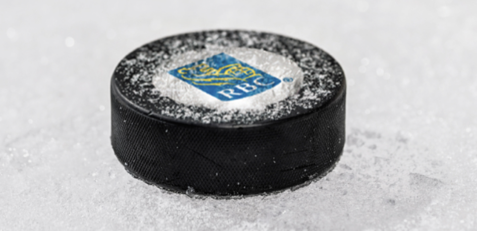 A black hockey puck with RBC logo on top, dusted with snow, rests on an icy surface. The scene conveys a cold, sporty atmosphere.