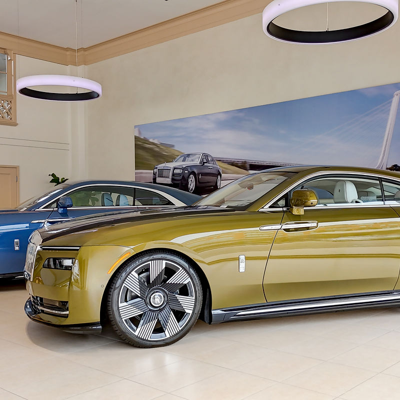 A gleaming olive green Rolls-Royce coupe is parked in a showroom next to a blue Rolls-Royce sedan. The cars are both modern luxury vehicles, with sleek designs and distinctive grilles. The showroom appears to be well-lit and spacious, with a large mural of Rolls-Royce cars in the background. Two circular pendant lights hang from the ceiling, adding to the modern aesthetic.