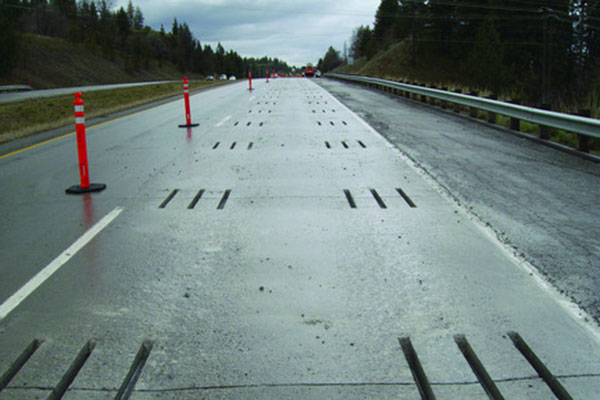 An elevated roadway view taken during active construction, looking down a highway lane that has been closed with orange and white vertical traffic delineator posts. Along the entire length of the center of the lane, numerous groups of parallel slots (ready for dowel bars) have been cut into the concrete surface. The image highlights the scale of traffic control required for a Dowel Bar Retrofit (DBR) project.