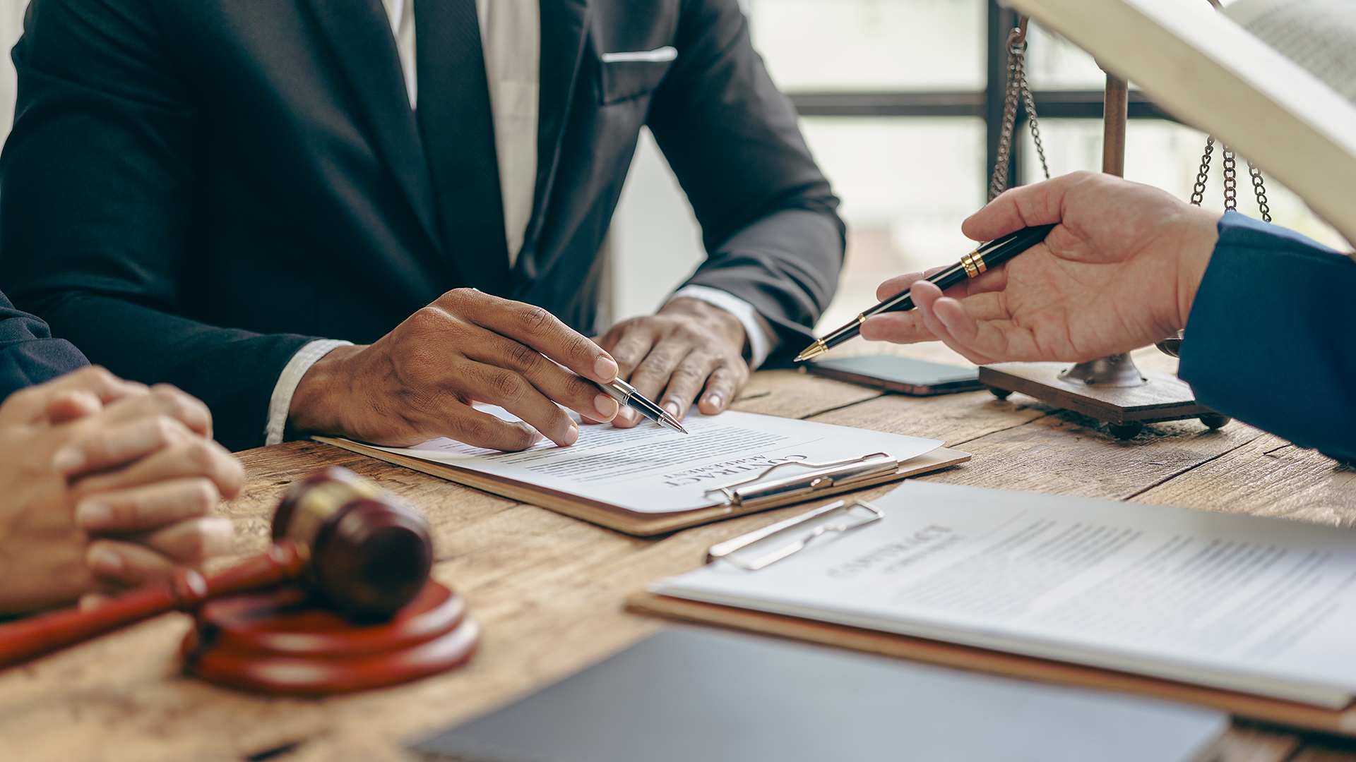 A close-up shot of three individuals in formal wear reviewing documents on a wooden desk. One person is pointing to a contract on a clipboard with a pen, while another person holds a pen and appears to be listening or about to sign. A gavel rests on the desk, along with a scale of justice in the background, suggesting a legal or business meeting setting. The lighting is warm and professional, focusing on the hands and documents.