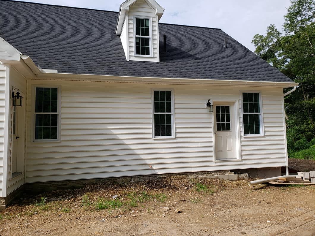 A newly built house exterior with white vinyl siding, black shingle roof, and a centered back door, featuring multiple windows, a small dormer, and unfinished ground surrounding the structure.