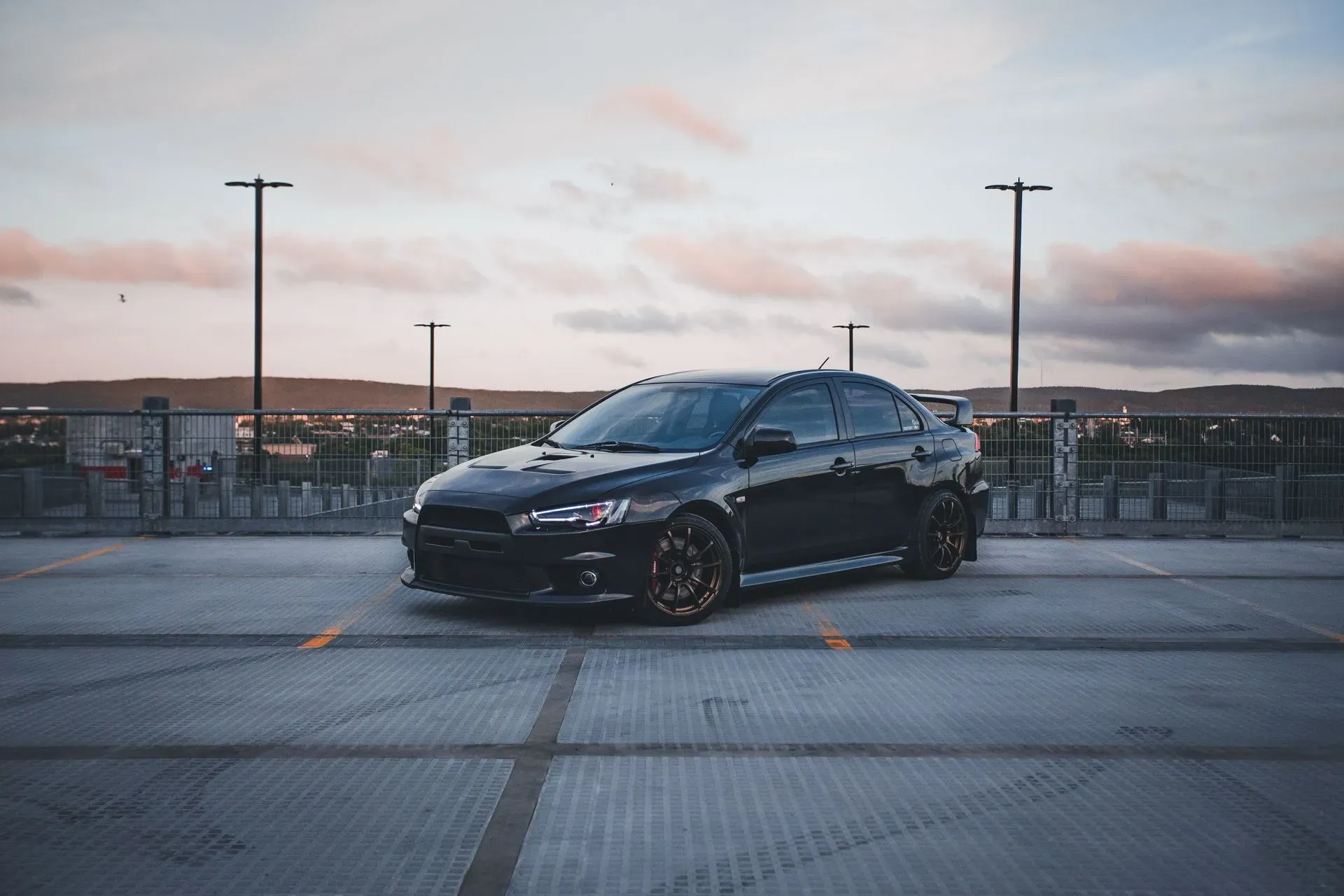 A black Mitsubishi Lancer Evolution X sedan is parked in a parking lot with a fenced overlook to a city and hills in the background. The sky is overcast with soft pink and orange hues of sunset. The car has a spoiler and aftermarket wheels.