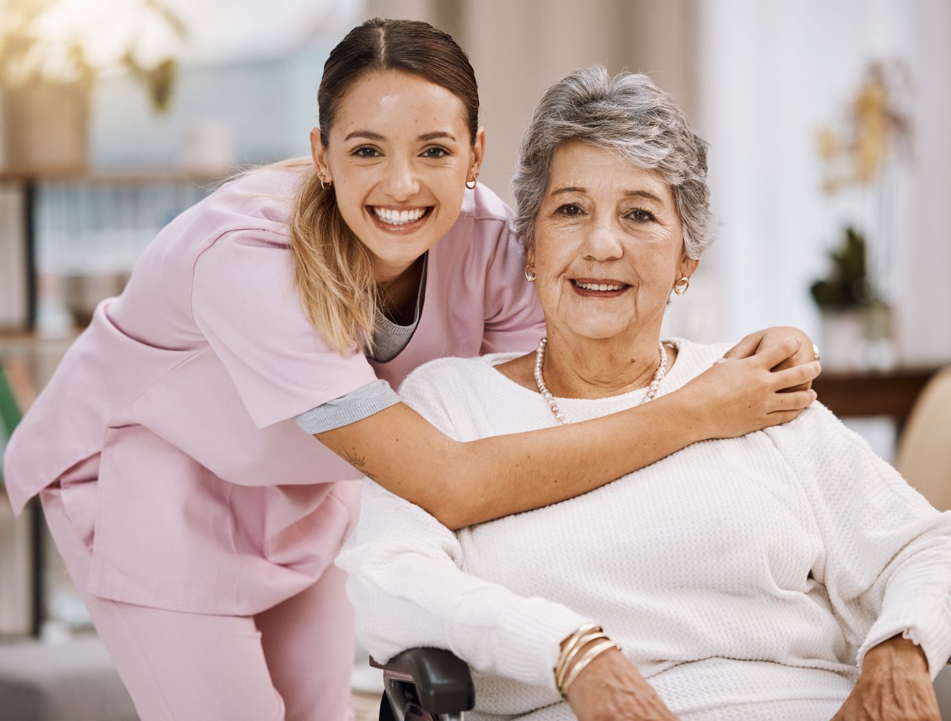 A young woman in pink scrubs embraces an elderly woman seated in a wheelchair, both smiling warmly at the camera. The scene appears to be indoors, possibly in a home or care facility, with soft lighting and a blurred background suggesting a cozy and supportive environment. The younger woman has her arm around the elder's shoulders, and the elder woman wears a pearl necklace and a white sweater.