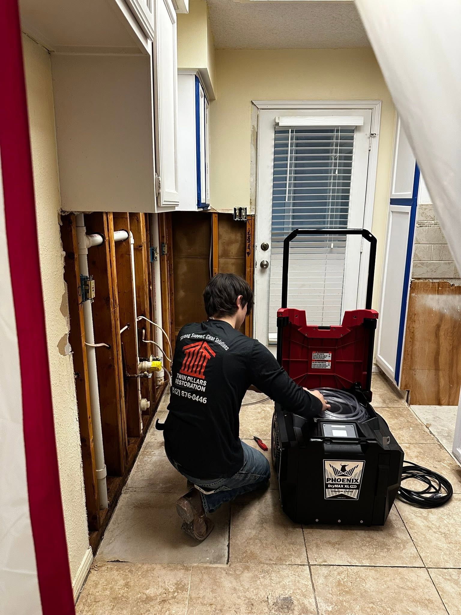 A Twin Pillars Restoration worker prepares drying equipment beside exposed wall framing and plumbing 