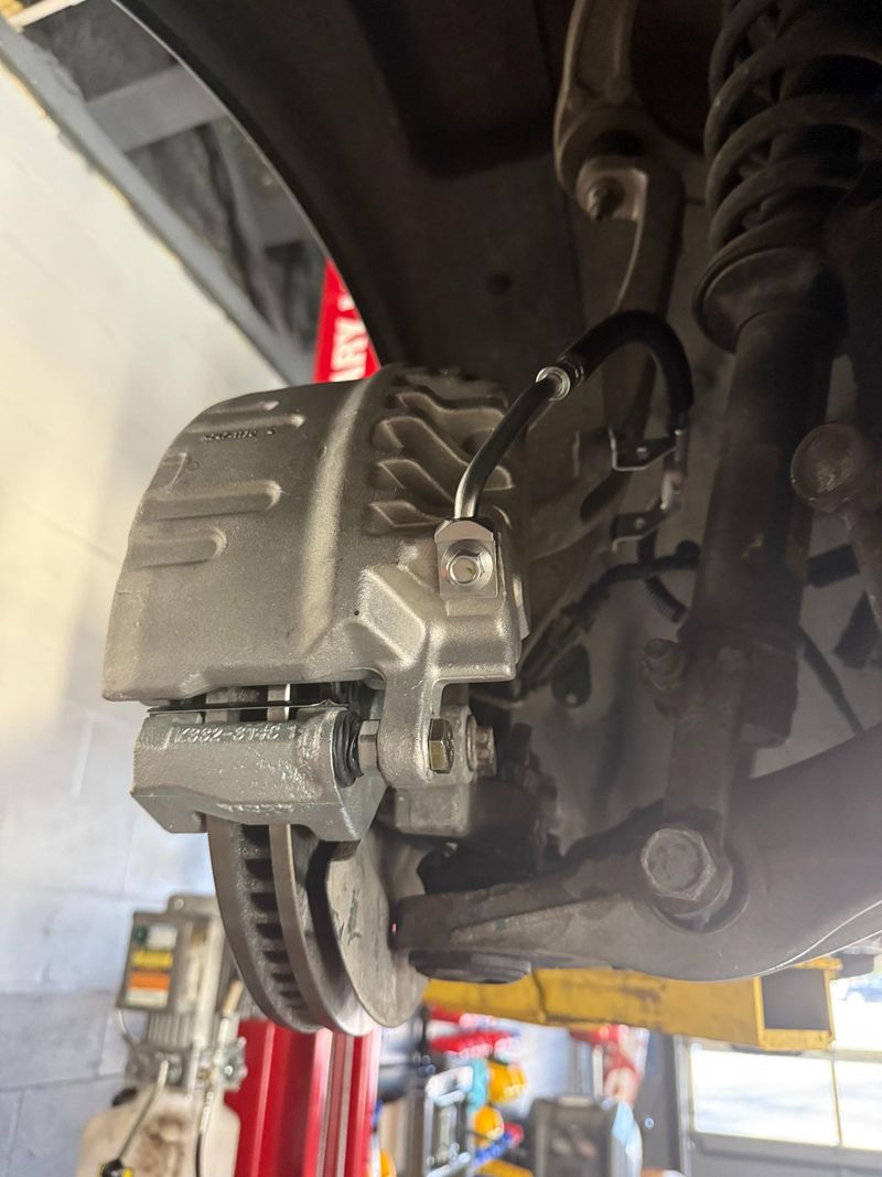 This is a close-up image of a vehicle's undercarriage showing a brand new, silver brake caliper and rotor assembly, professionally installed and connected to the brake line, suggesting recent brake service.