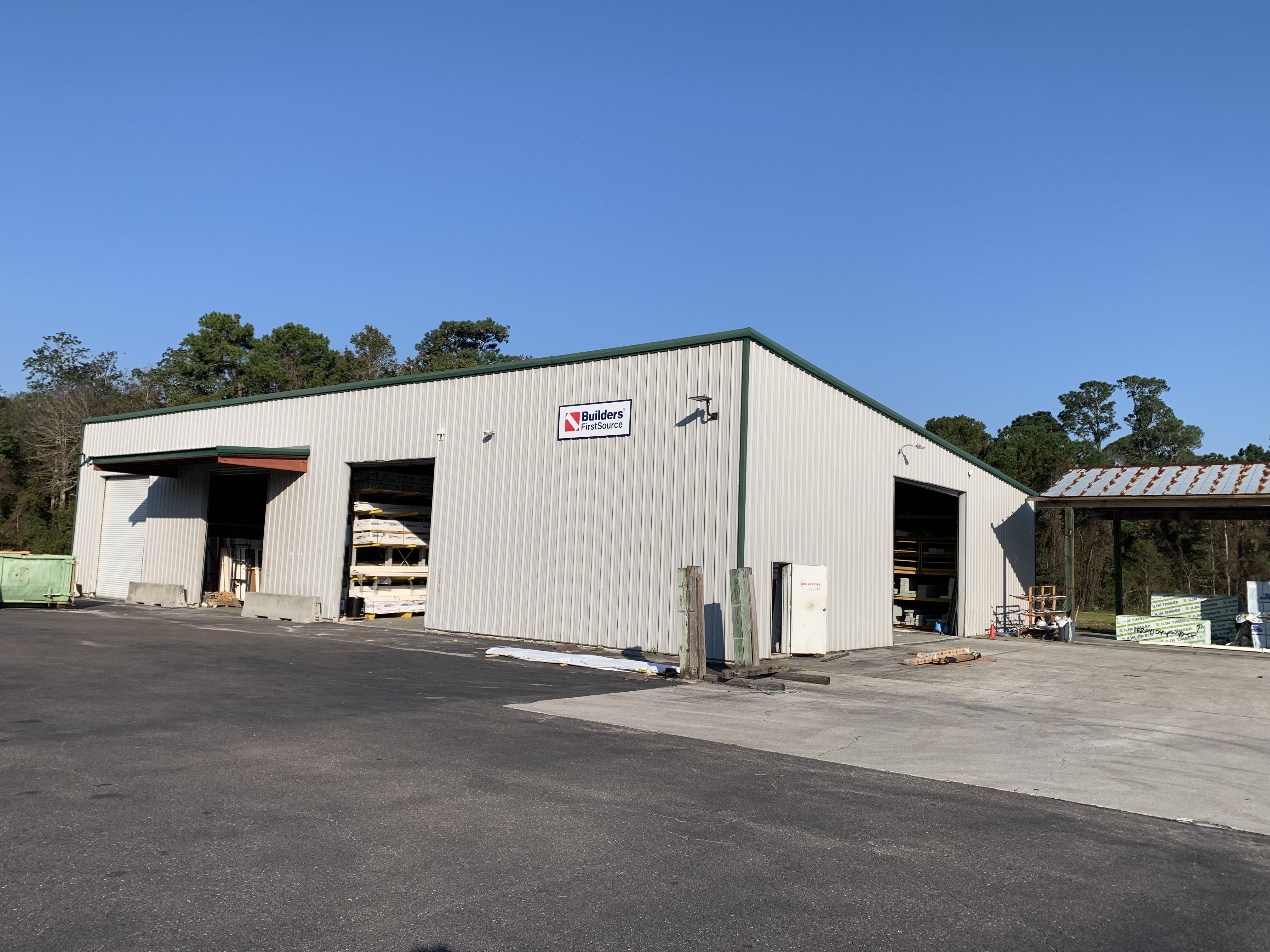 A large metal warehouse building with two open bays is visible. The building has beige siding and a green roof, with stacks of building materials like wood and other supplies stored inside. A sign on the side of the building reads "Builders FirstSource." The area outside has a large paved surface, with some additional materials and a dumpster placed nearby. Trees are visible in the background under a clear blue sky.