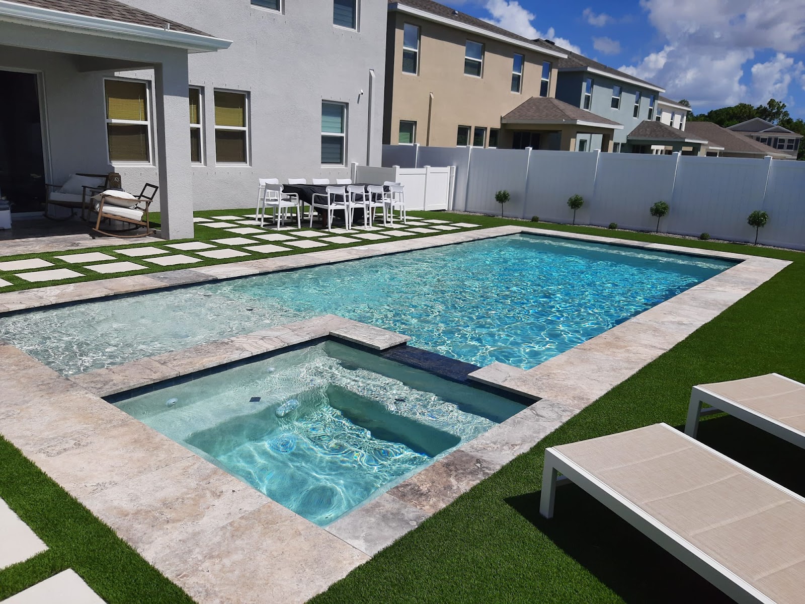 Modern backyard swimming pool with an attached raised spa, light stone pavers, artificial turf, and lounge seating.
