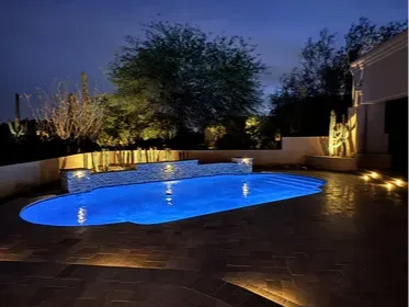 Scottsdale pool at night with LED lighting, stone water feature wall, and travertine decking. The uplighting on the desert landscaping is doing a lot of work here, giving the whole yard a resort feel without a single fire feature in sight. Built by Hacienda Outdoors, ROC #325952.