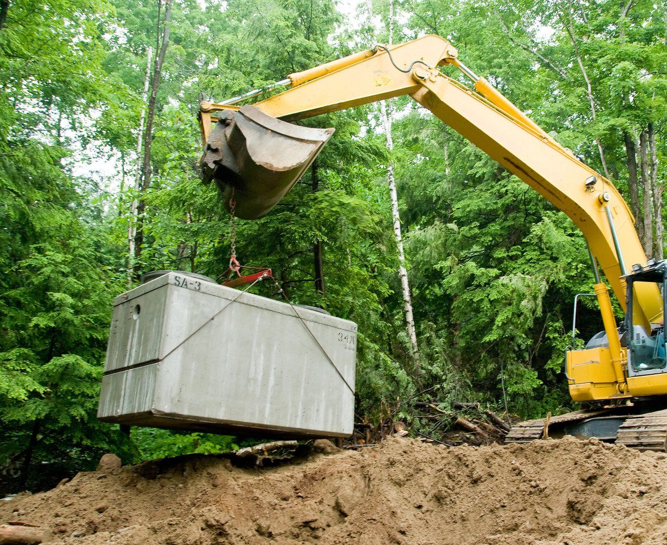 A yellow excavator uses a heavy-duty chain to lower a massive precast concrete septic tank into position. The installation takes place at a wooded construction site with fresh dirt mounds.