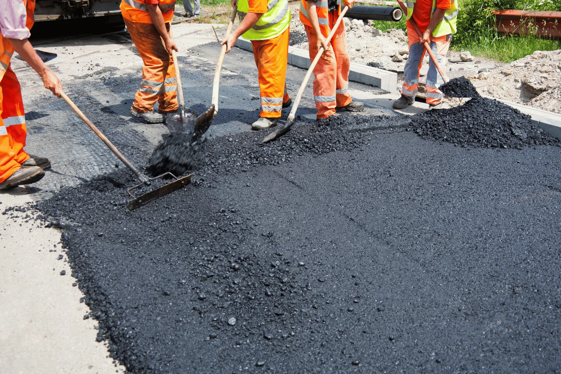 A group of construction workers in orange safety vests and pants are spreading fresh asphalt on a road. Some workers are using shovels to move the hot asphalt, while others are using rakes to level it. The ground is covered in dark grey asphalt with some lighter grey concrete on the side. There is some greenery and construction materials visible in the background.