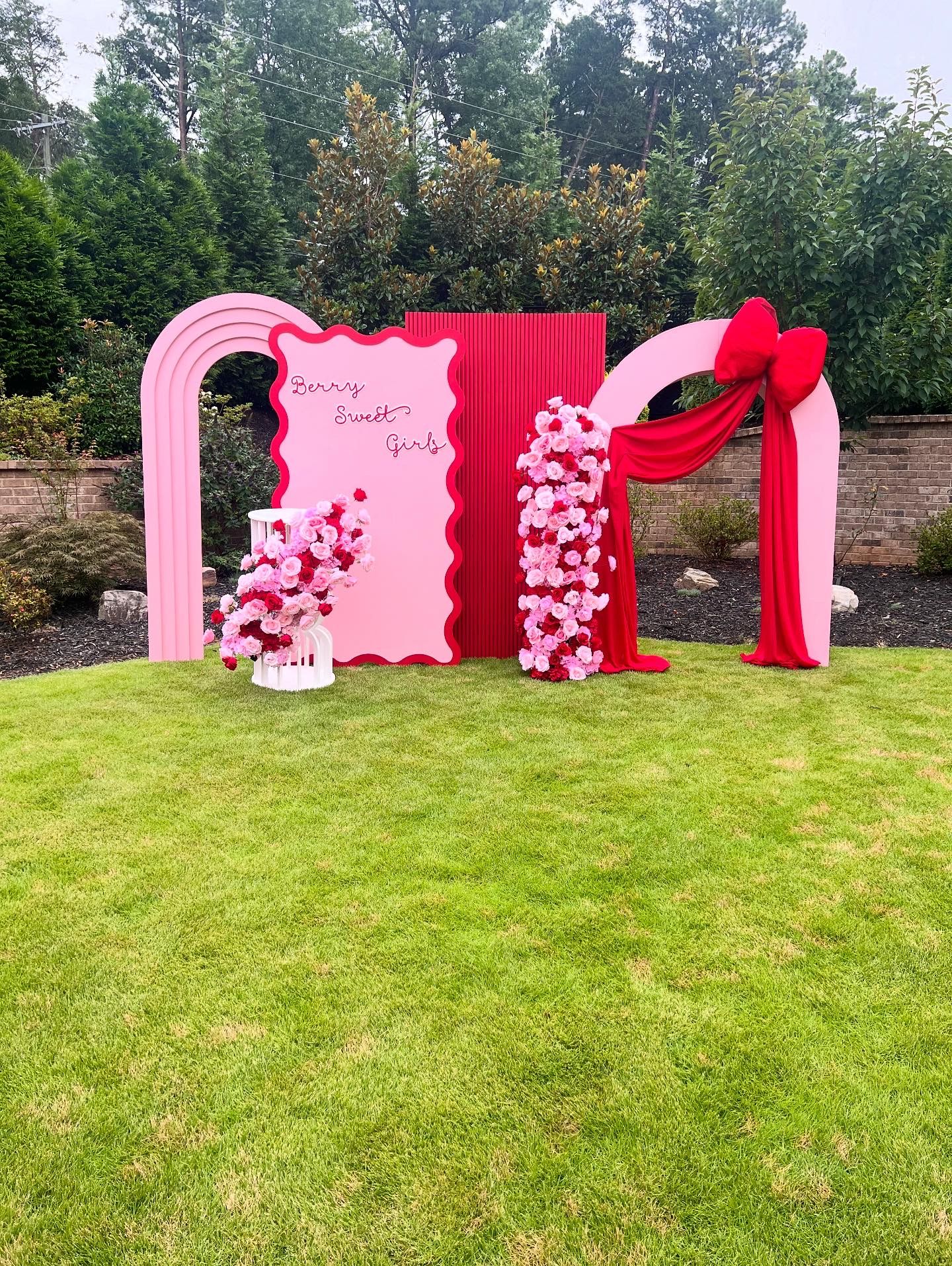 Outdoor event backdrop with pink and red arched panels, floral arrangements, draped fabric bow, and &ldquo;Berry Sweet Girl&rdquo; sign, styled on green lawn for a charming, elegant celebration setup.