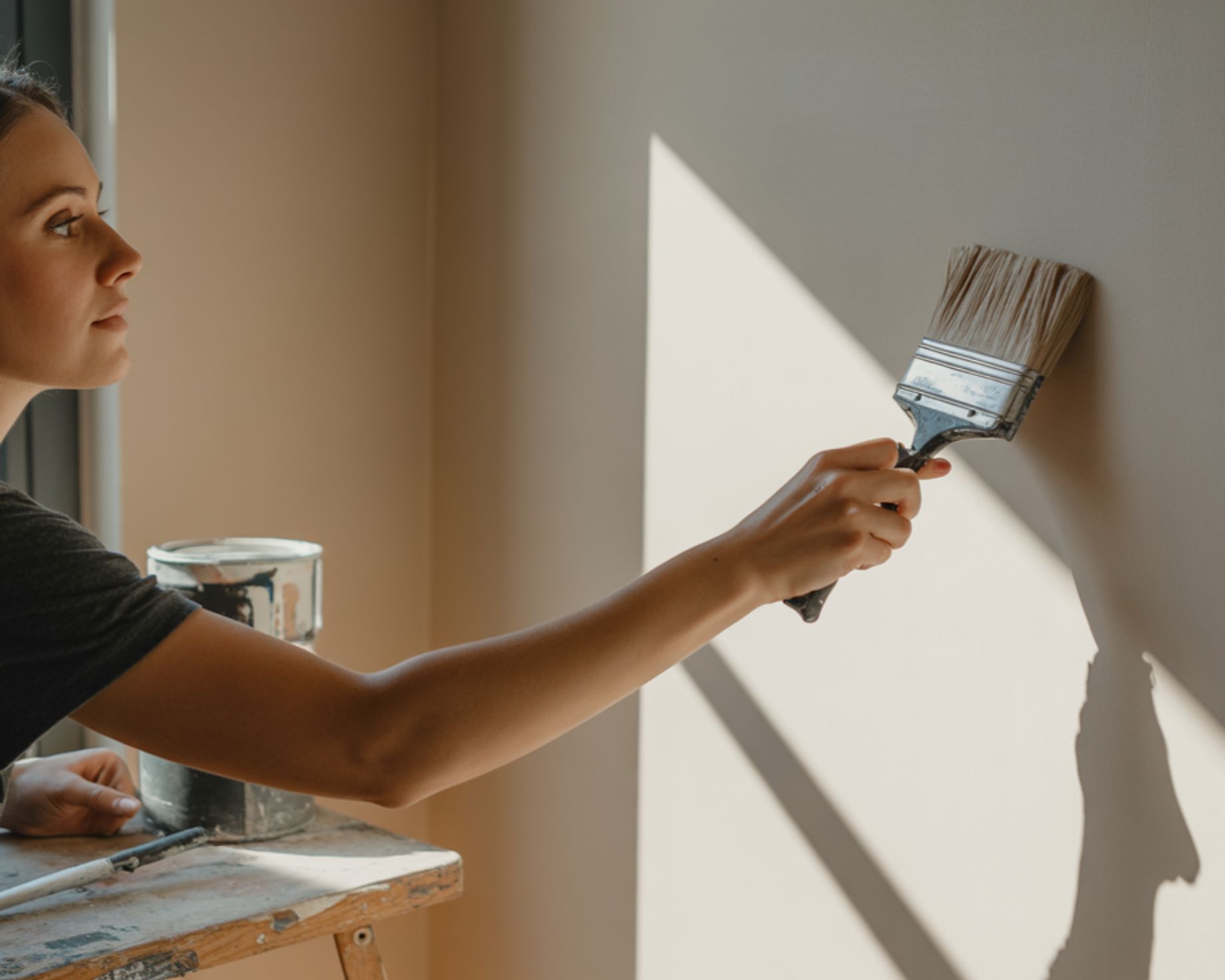 A woman is painting a wall with a paintbrush, with a can of paint on a step stool next to her. Sunli 