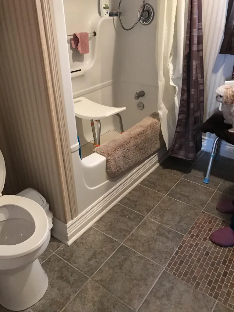 A bathroom with a toilet, bathtub, shower seat, and a small dog. The bathroom has tiled floors and a shower curtain. There are also two pairs of slippers on the floor.