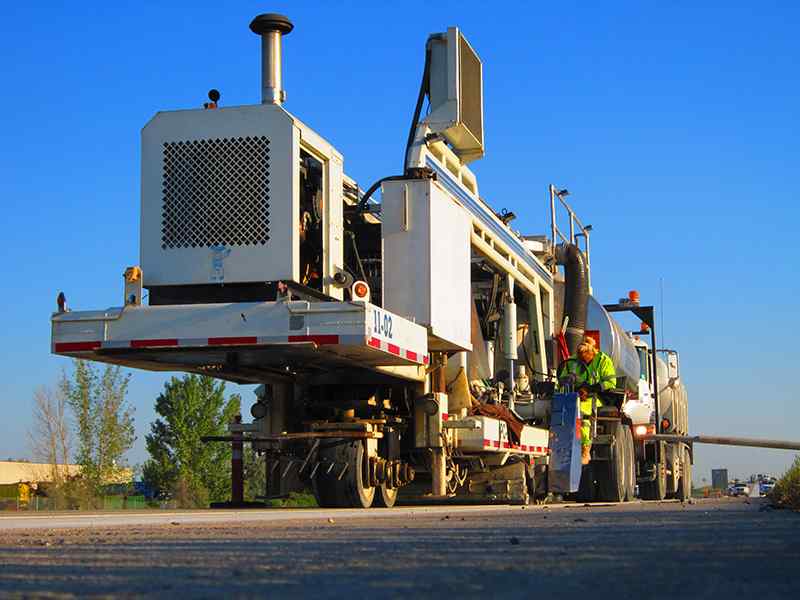 Low-perspective, ground-level photograph of the front chassis and heavy-duty wheels of a DSI diamond grinding machine staged on concrete. An operator in a high-visibility jacket sits at the side control station under a bright, clear blue sky.