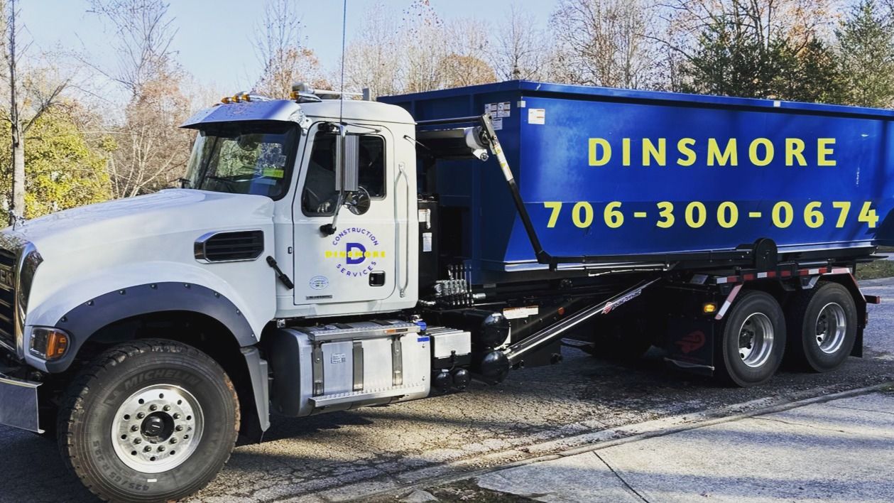 A large white dump truck with a blue container labeled Dinsmore and a phone number is parked outdoors along a roadside with trees in the background.