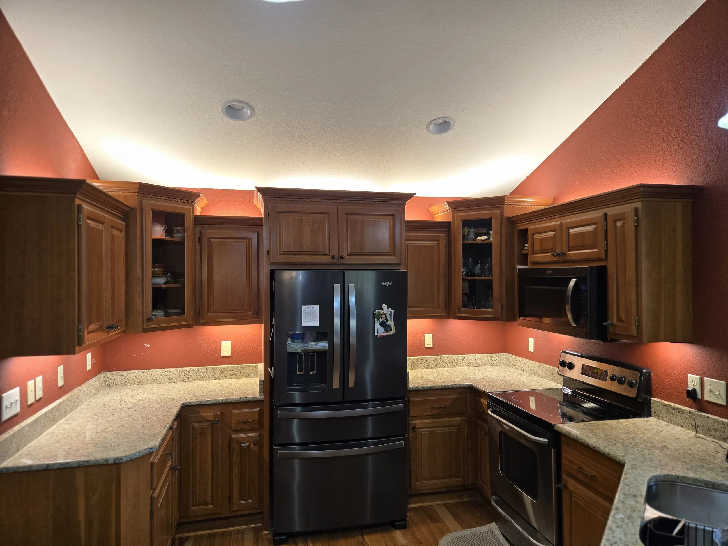 A modern kitchen with dark wood cabinets, granite countertops, and a red accent wall is showcased. A large stainless steel refrigerator with a water and ice dispenser is the central appliance. To the right of the refrigerator, there's a built-in microwave above a stovetop, and to the far right, a stove with a digital display. The kitchen is illuminated by recessed ceiling lights and subtle LED strip lighting under the cabinets, creating a warm ambiance.