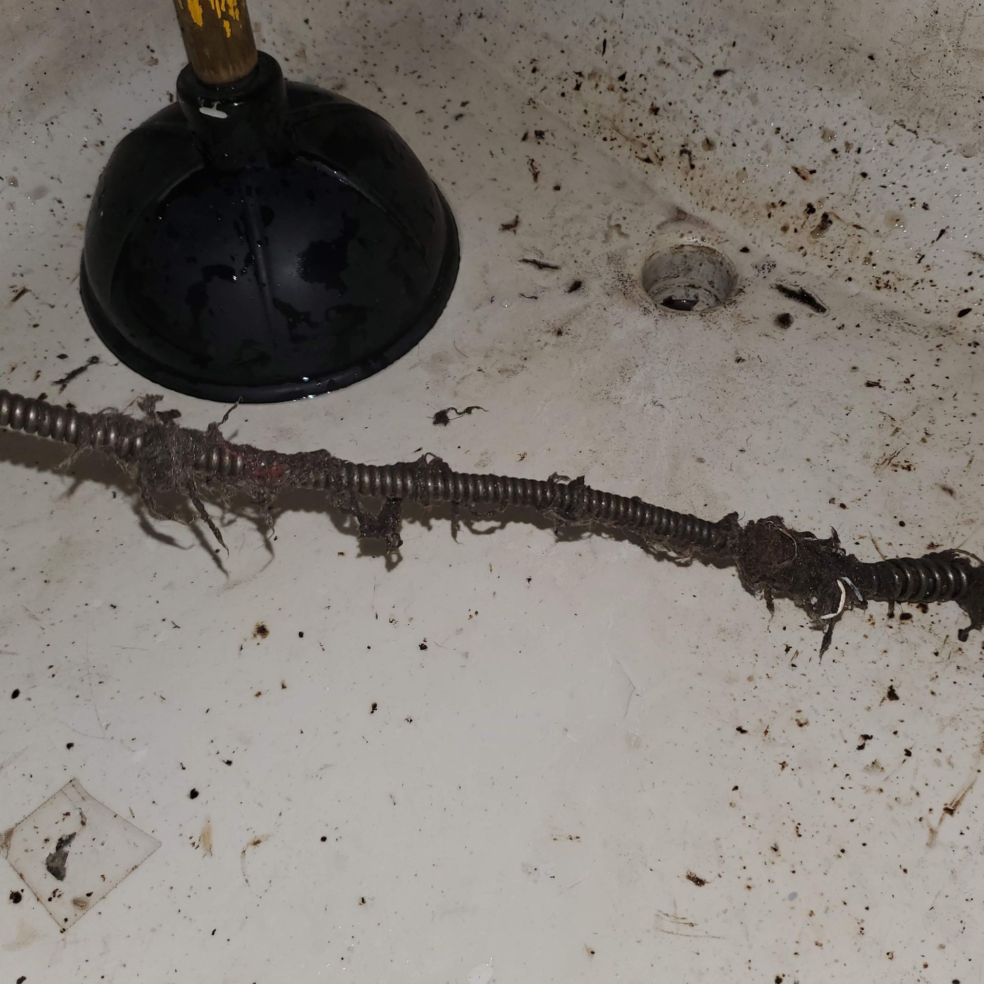 A dirty sink surface with a plunger and drain opening visible, alongside a drain snake covered in grime, hair, and debris, showing a recently cleared clog and messy buildup removal.