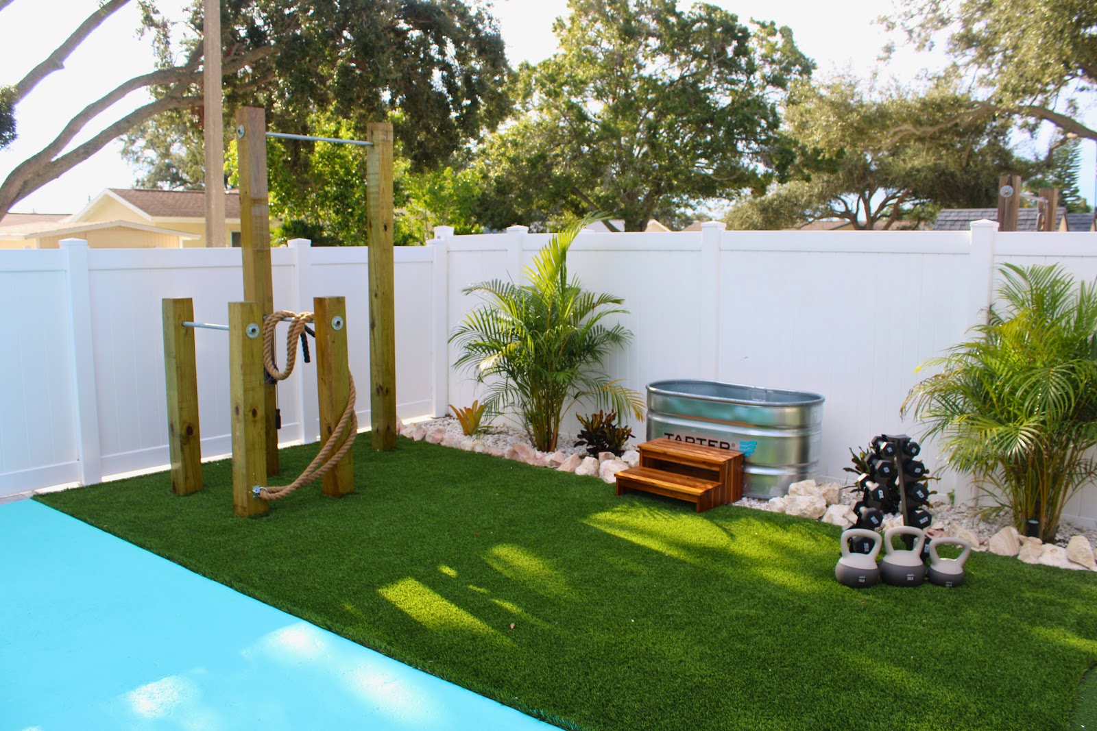 Backyard poolside area with artificial turf, outdoor fitness equipment, tropical plants, and a white privacy fence.