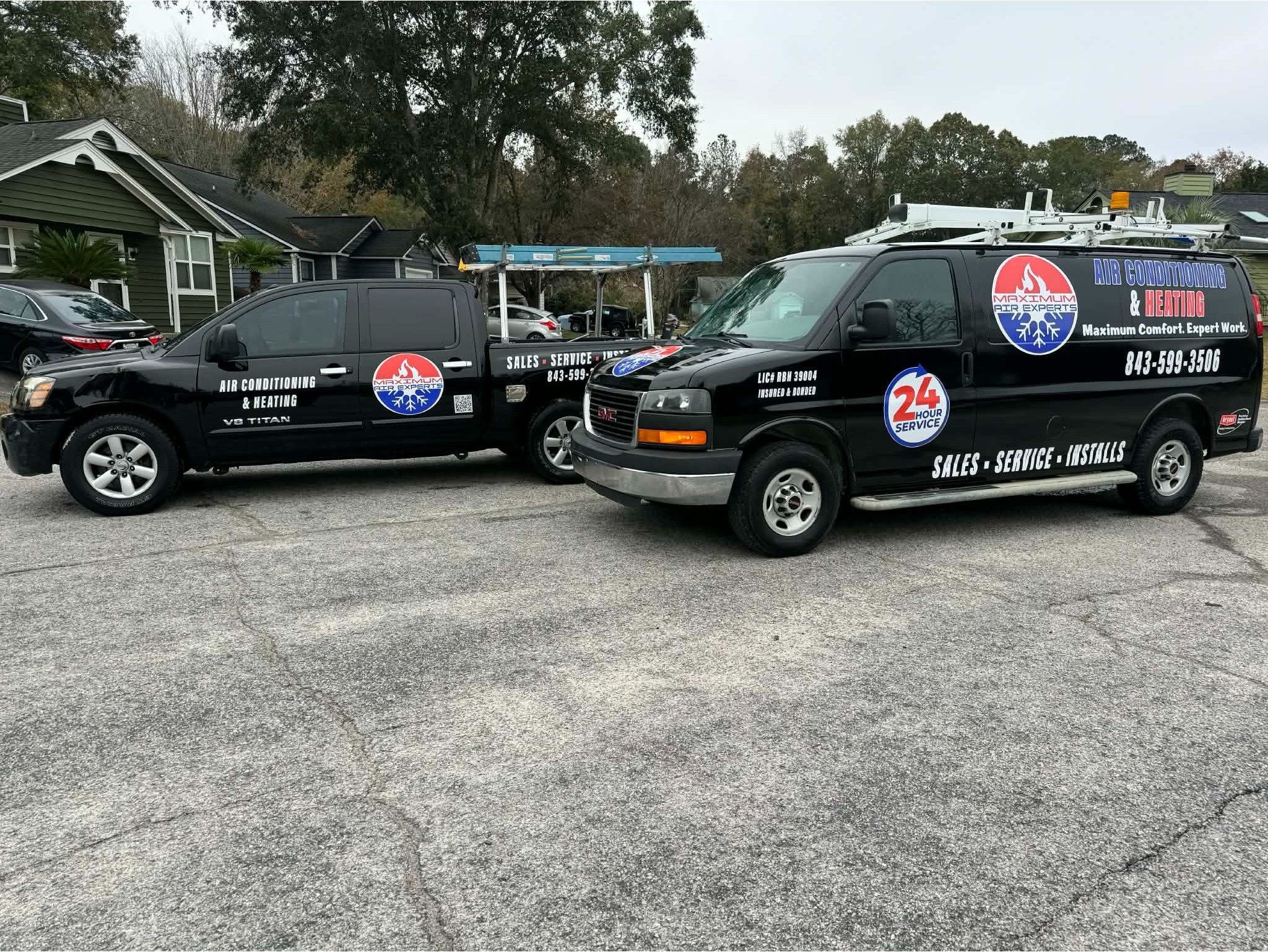 Two black Maximum Air Experts service vehicles&mdash;a Nissan pickup truck and a GMC work van equipped with roof ladders&mdash;parked on a paved driveway in a residential neighborhood. Both vehicles feature prominent branding, the company logo, contact information, and decals reading "Air Conditioning & Heating" and "24 Hour Service."