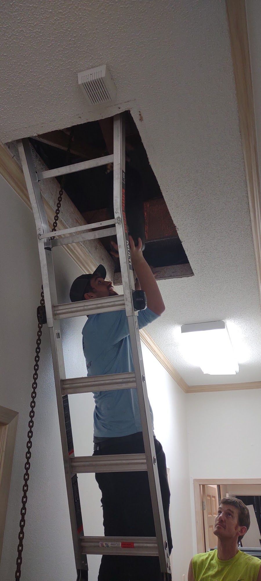 Technician standing on ladder accessing ceiling opening to inspect or repair attic HVAC ductwork, while another person observes below in indoor residential setting with lighting fixture and ceiling vent.