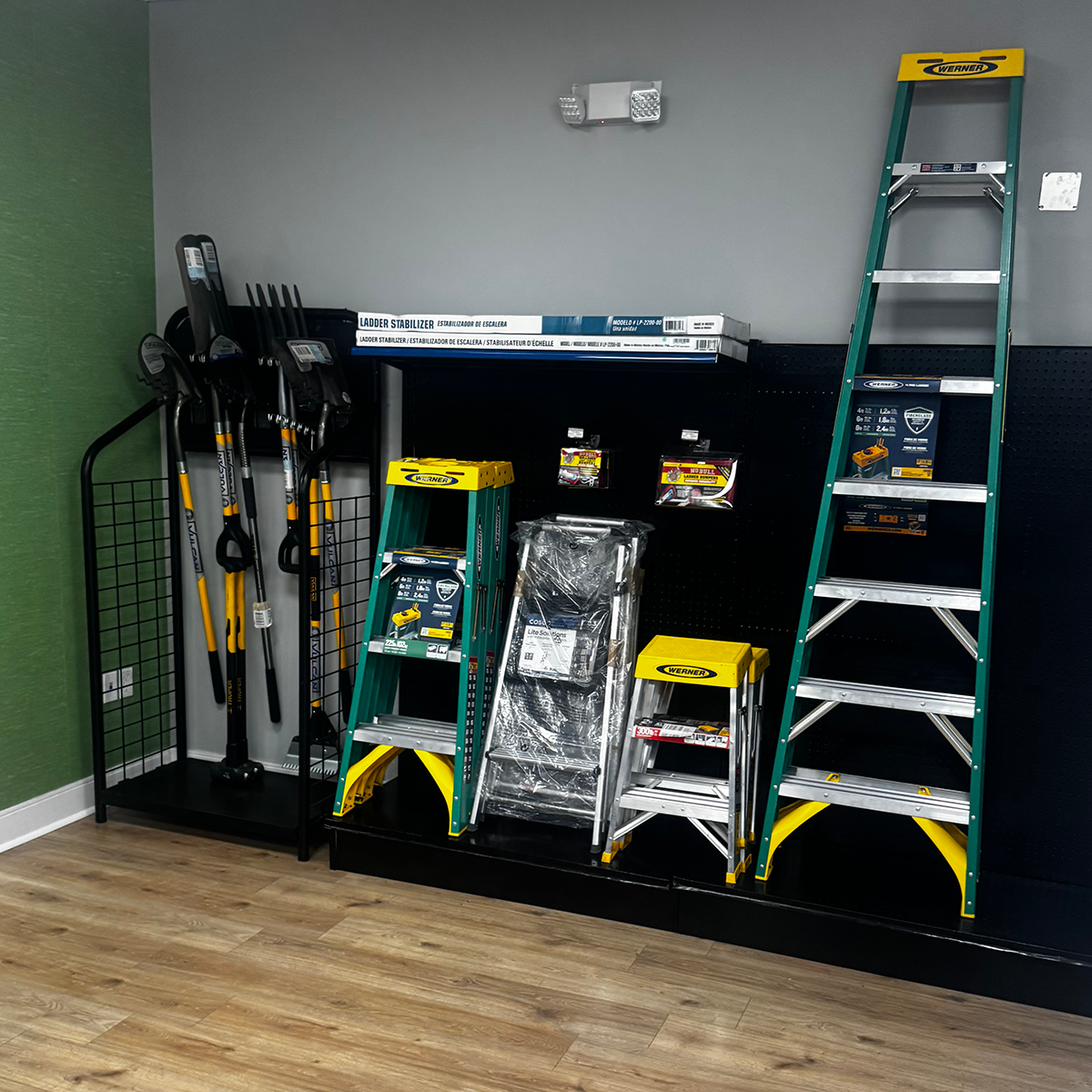 Builders FirstSource display featuring green-and-yellow Werner step ladders and an extension ladder on a black platform. Shovels and other long-handled yard tools stand in a rack at left, with packaged ladder accessories and stabilizers arranged on the wall behind them.