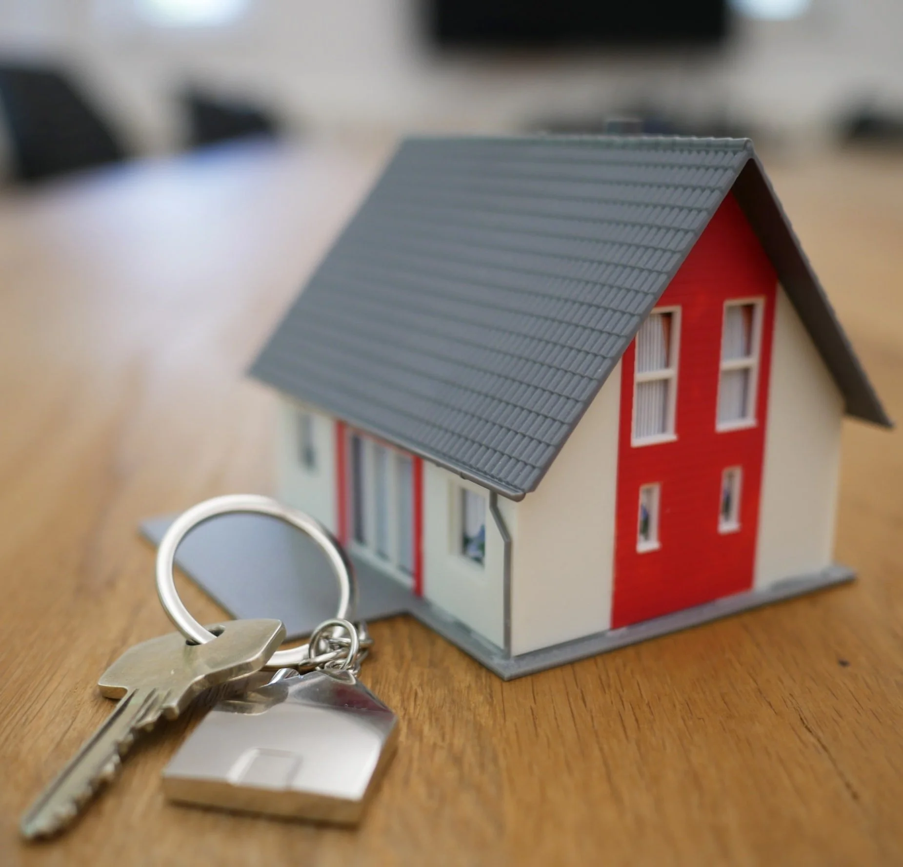 A close-up shot shows a miniature house model with a red facade and white windows, sitting on a wooden surface. In the foreground, a silver key is attached to a keyring with a house-shaped charm, suggesting concepts of homeownership or real estate.
