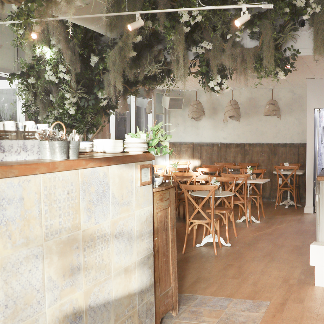 A dining room layered with flowering vines, textured greenery, warm wood, and soft light creates the feeling of sitting beneath a garden canopy.