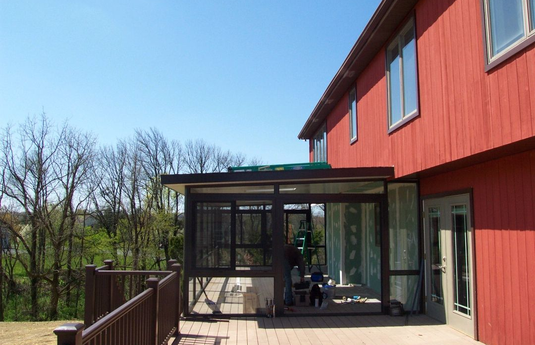 Modern home addition featuring a partially enclosed patio with dark framing and large screened panels. Attached to a red exterior house, the structure is under construction, offering an extended outdoor living area with scenic backyard views and natural light.
