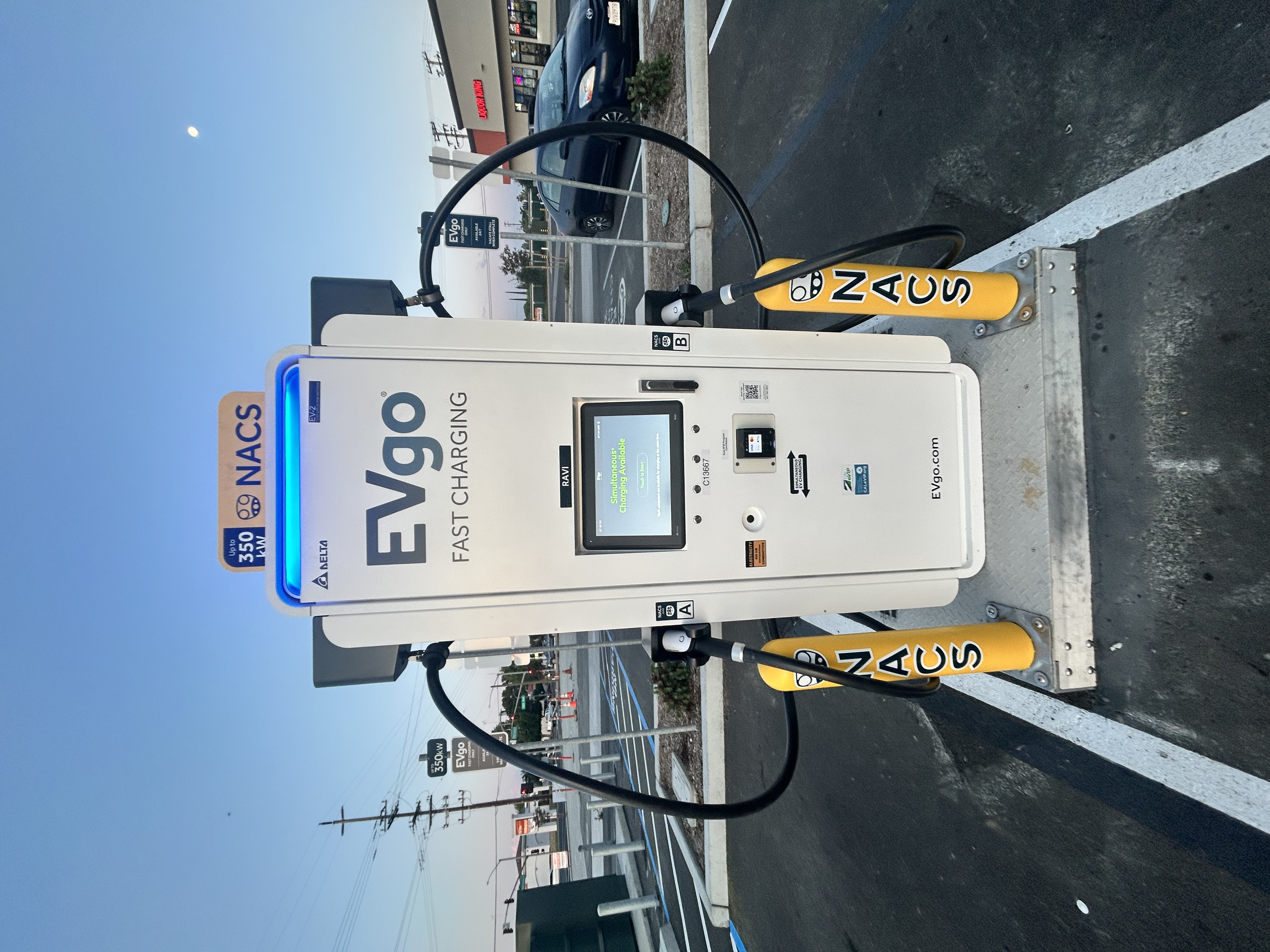 NACS EVgo fast charging unit installed in marked parking space at a public charging station, with EVgo logos on the charger, and yellow protective bollards around the unit