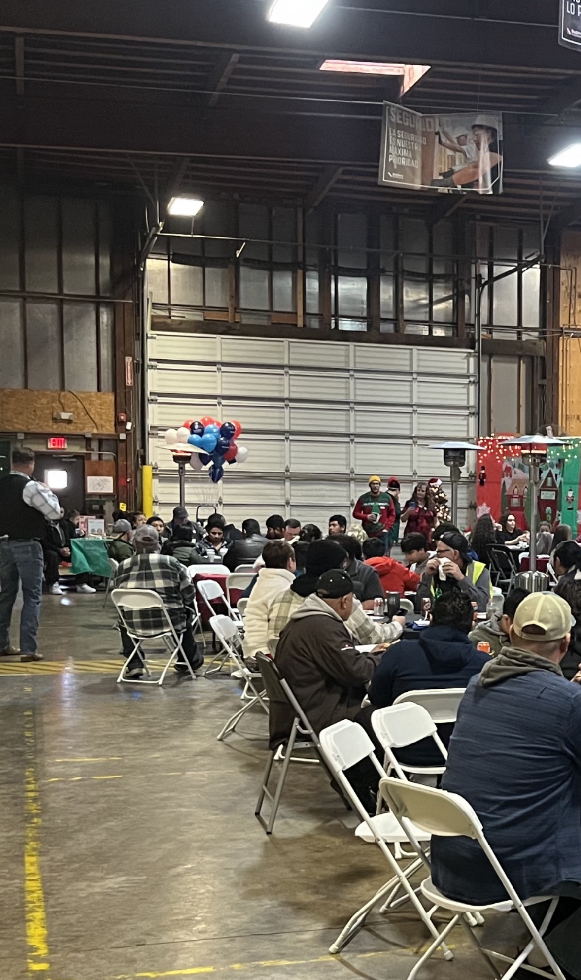 A large group of people gathered in an industrial warehouse for an indoor event. The room is filled with rows of folding chairs and tables, where attendees are seated, eating, and socializing. In the background, a couple of people wearing festive holiday outfits stand near red, white, and blue balloons. The walls are lined with metal panels, and overhead lights provide bright illumination. There is also holiday-themed decor, including lights and a small Christmas tree, adding a festive atmosphere to the gathering. A large garage-style door is visible on the far side of the room.