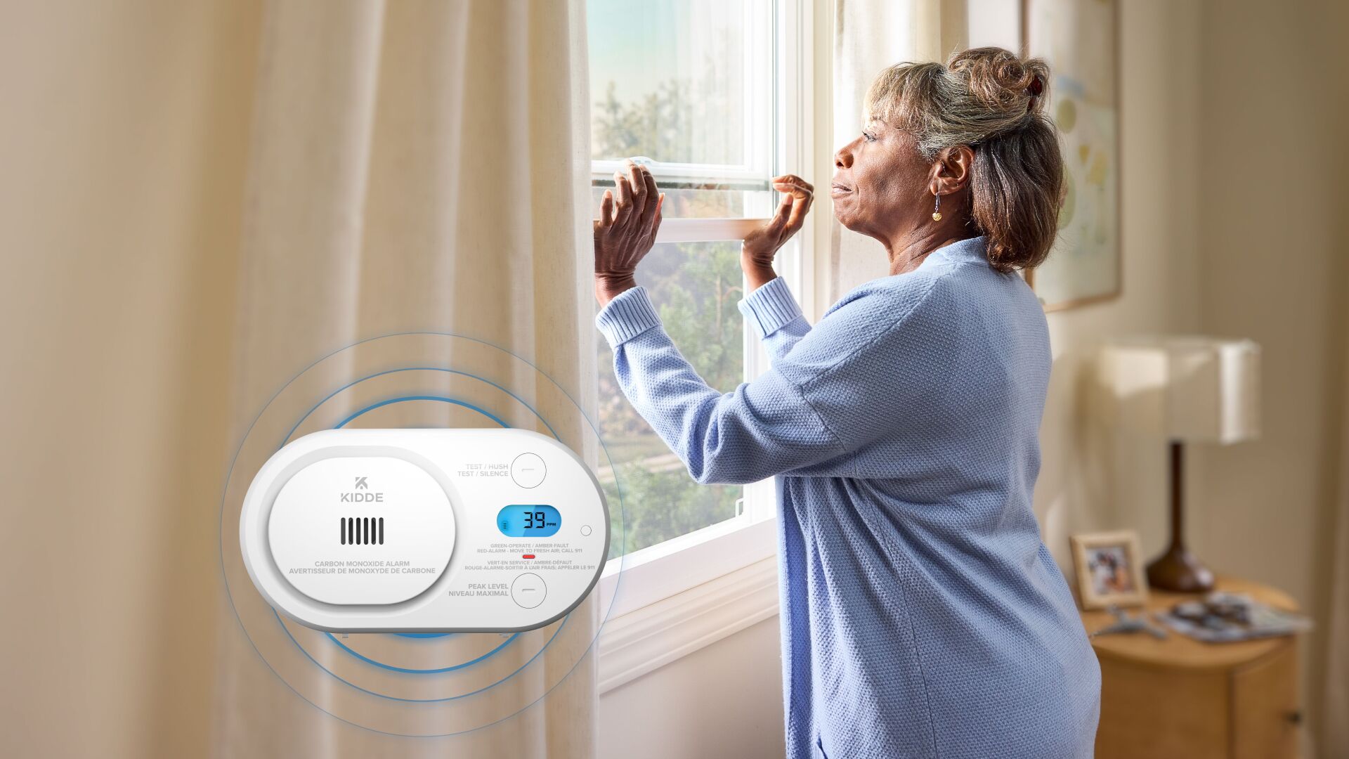 A woman opens her window. In the foreground, a carbon monoxide alarm is going off, showing that 39 ppm has been detected in the air. Blue circles representing the sound is emanating from the device.