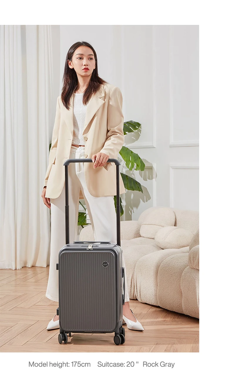 A woman in a beige blazer and white pants stands with a dark gray suitcase in a well-lit room. She is holding the suitcase's handle and looking off to the side. The background features cream-colored curtains and a textured sofa, with a large green plant visible on the right. The floor appears to be made of light wood.