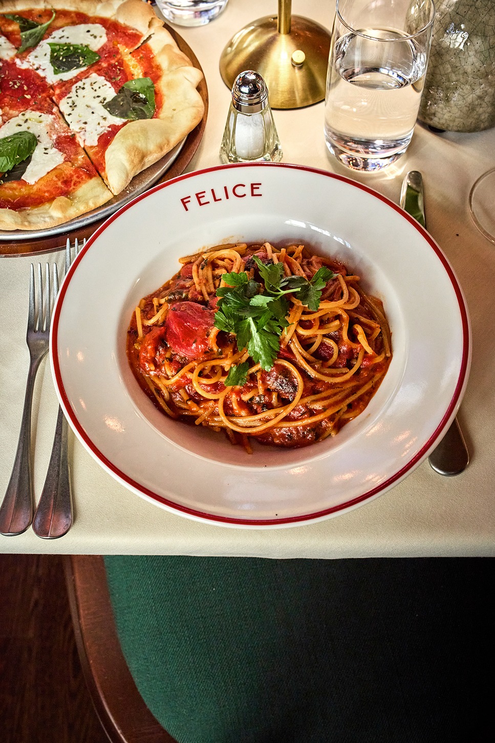 A top-down view of a classic spaghetti pomodoro served in a white bowl. The pasta is coated in a rich red sauce and topped with a sprig of fresh basil, set against a clean white tablecloth.