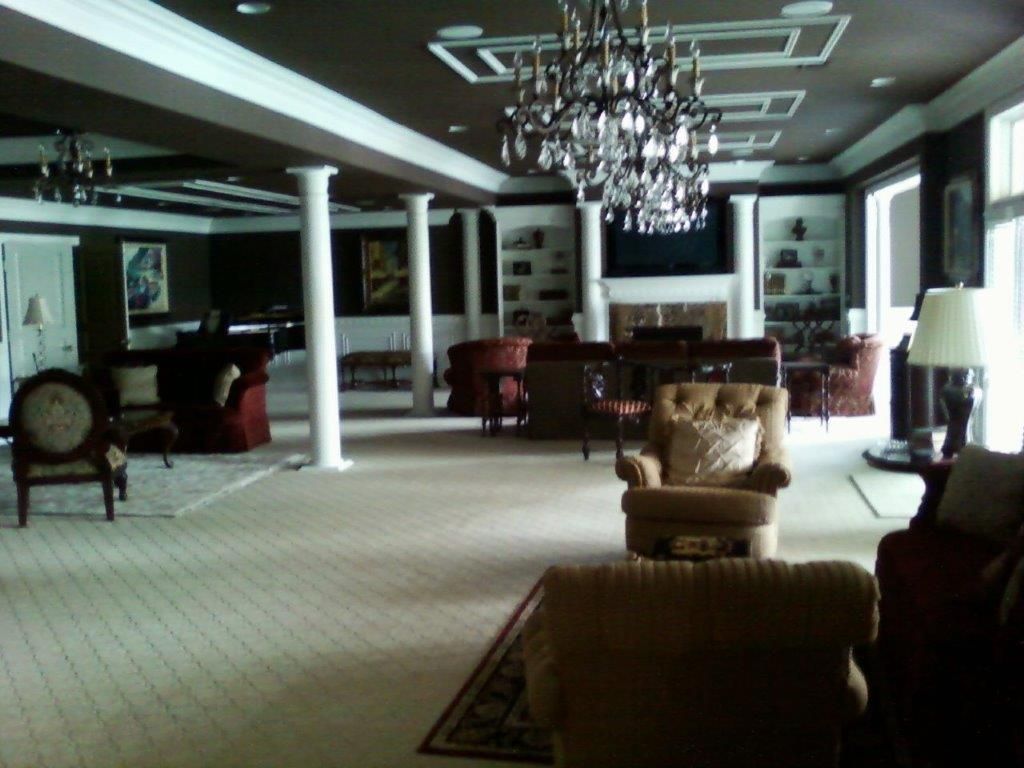 A spacious, elegant living room with high ceilings, decorative columns, and a large chandelier. Multiple seating areas, built-in shelves, and a fireplace create a luxurious and comfortable gathering space.