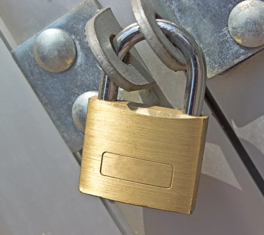 A close-up shot of a brass padlock securing a metal latch. The padlock has a textured surface and a rectangular indentation. The latch is attached to a grey surface with visible rivets. A dark cable is partially visible to the right of the padlock.