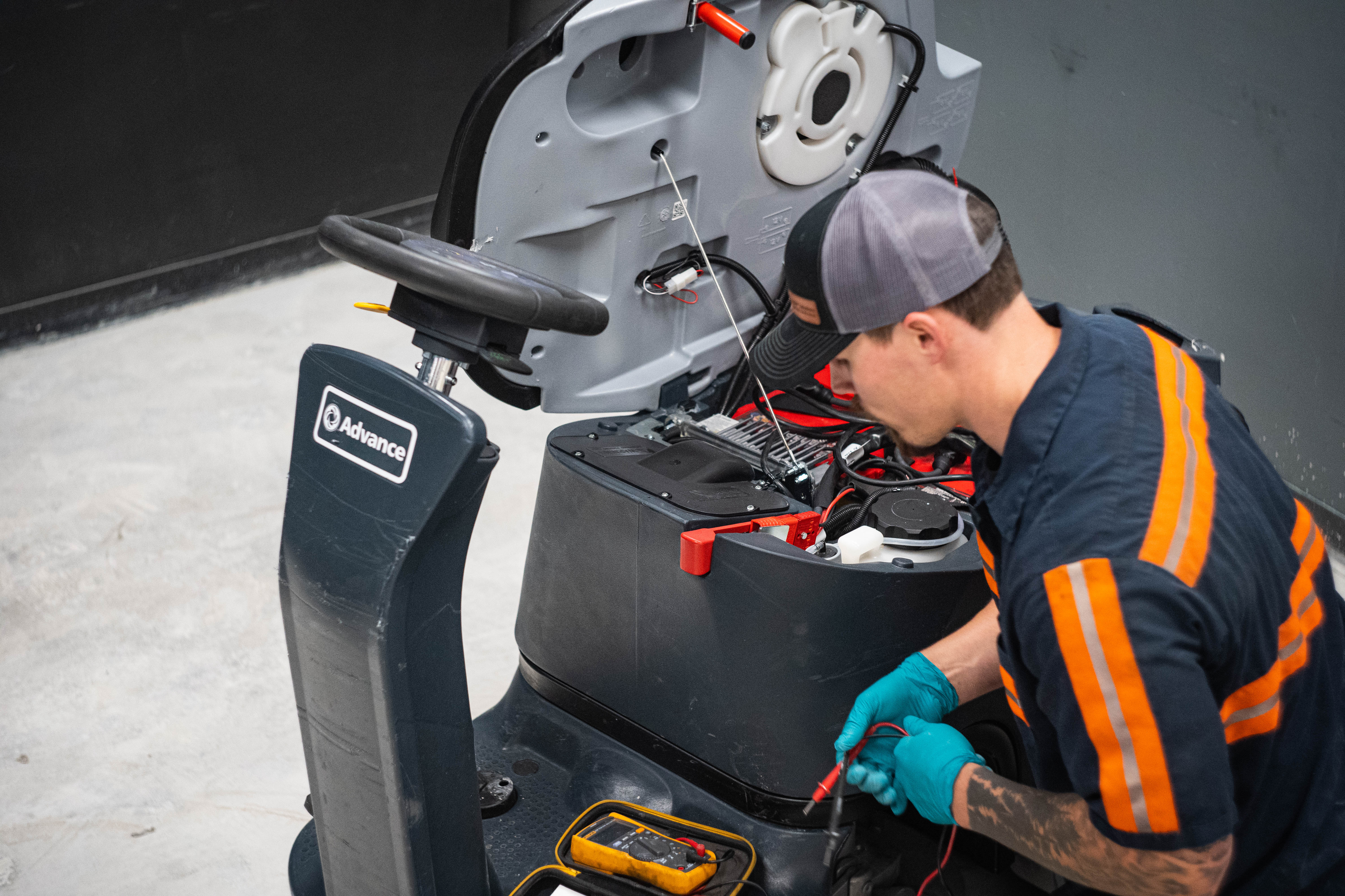 An expert technician at WaySolutions utilizes a digital multimeter to test the complex electrical system inside an Advance ride-on commercial floor scrubber. With the equipment's housing opened to reveal the deep-cycle batteries and wiring harness, this image highlights our commitment to data-driven diagnostics and high-level electrical repair for heavy-duty industrial cleaning fleets.