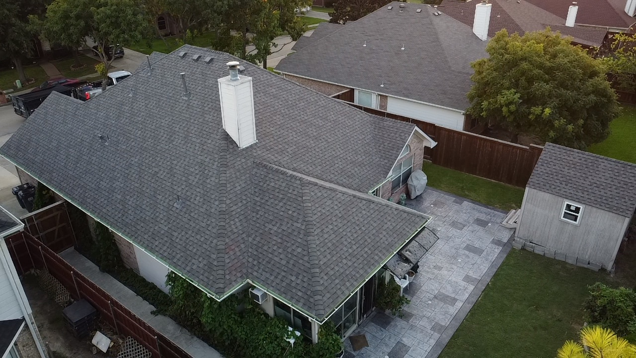 An aerial view captures a suburban neighborhood with multiple houses featuring dark gray asphalt shingle roofs. One prominent house in the foreground has a white chimney and a large, grey, textured patio. Adjacent to the patio is a small, light-colored shed with a single window. The properties are surrounded by green lawns, trees, and wooden fences. Several vehicles, including trucks and a dumpster, are visible on the street in the background.