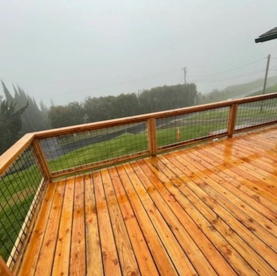 A newly installed wooden deck with a clear protective finish, appearing wet from the rain. The railing features wooden posts and top rail with black wire mesh infill, overlooking a misty landscape.