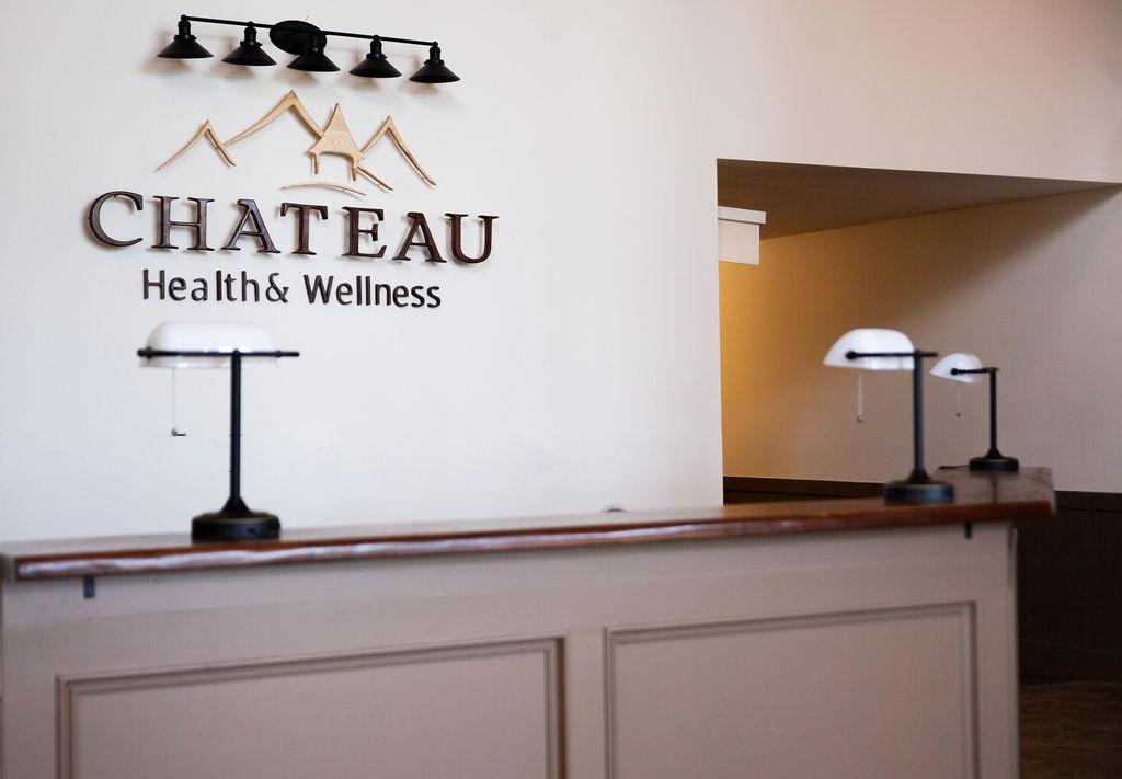 front desk lobby area with 3 lamps along an angled desk. To the left on the wall is a wooden carved logo with a mountain siloute above that says "Chateau Health & Wellness"