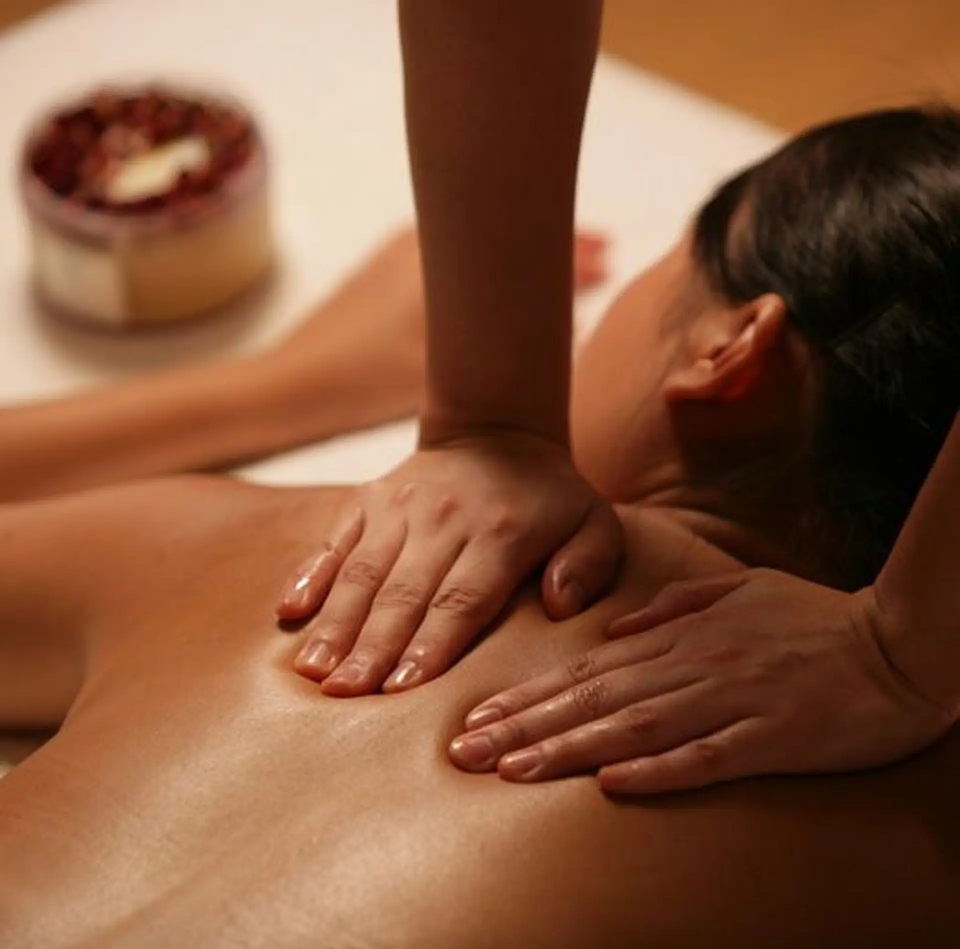 A close-up shot shows a person receiving a massage on their back. Two pairs of hands are visible, one pair working on the upper back and shoulders, and the other pair on the lower back. A blurred cylindrical container filled with what appears to be dried petals and a dollop of cream is visible in the background on the left. The lighting is warm and soft, highlighting the oils on the skin.