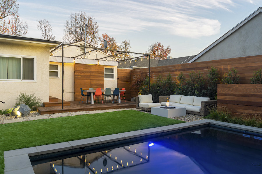 Modern backyard with blue-lit swimming pool, green lawn, wooden patio, outdoor dining and lounge seating, creating a warm, inviting retreat.