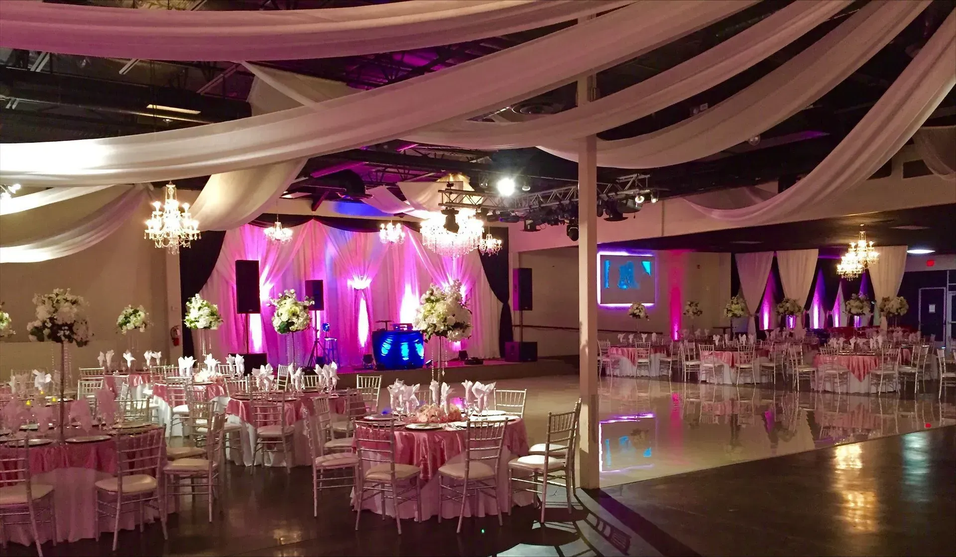 Elegant wedding venue featuring illuminated stage, crystal chandeliers, ceiling drapery, floral cent 