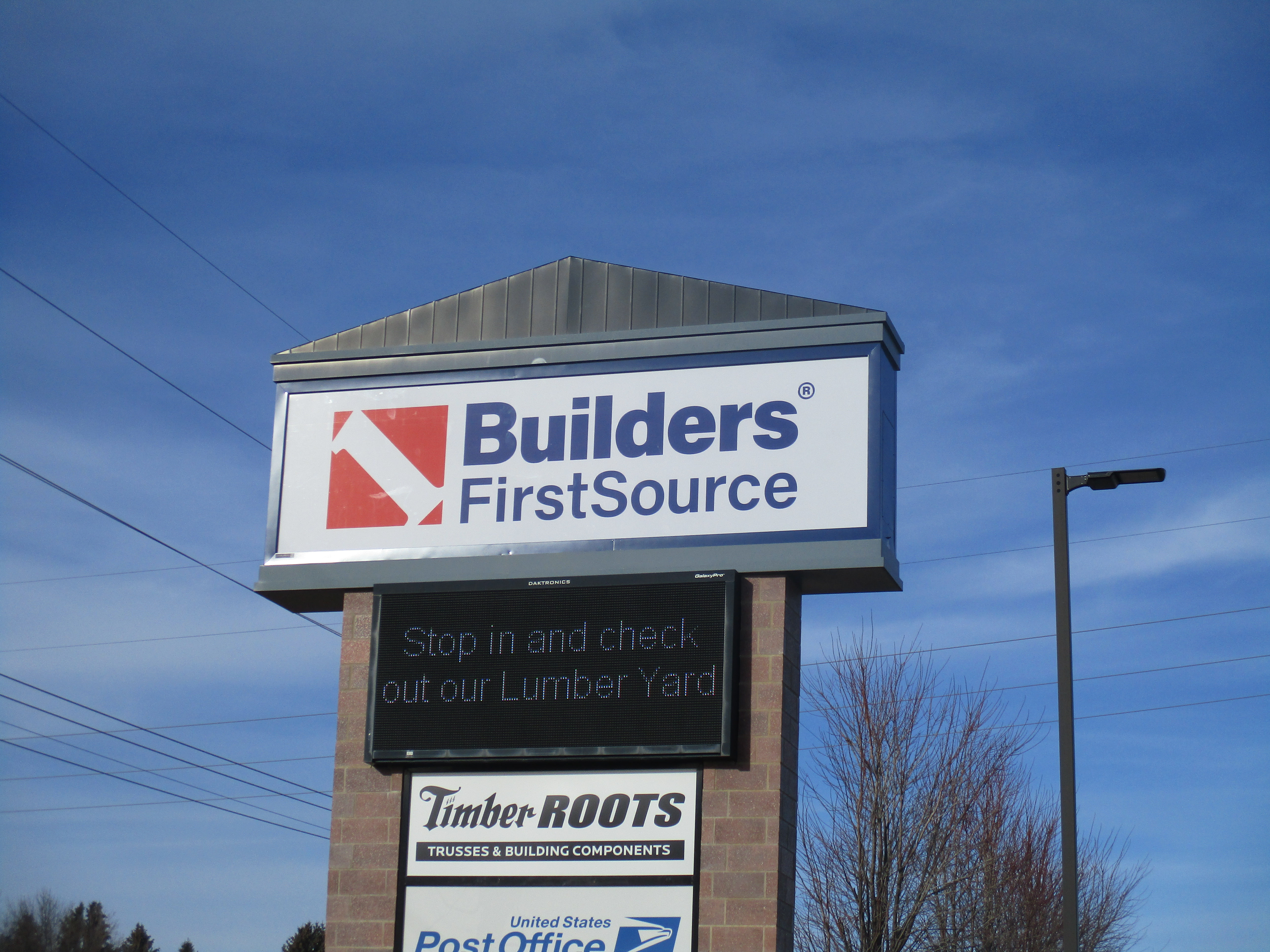 A roadside sign for Builders FirstSource, featuring a bold red and blue logo with "Builders FirstSource" prominently displayed. Below the main sign is an LED screen with the message "Stop in and check out our Lumber Yard" and a sign for "Timber Roots Trusses & Building Components." The sign is mounted on a brick base under a clear blue sky.